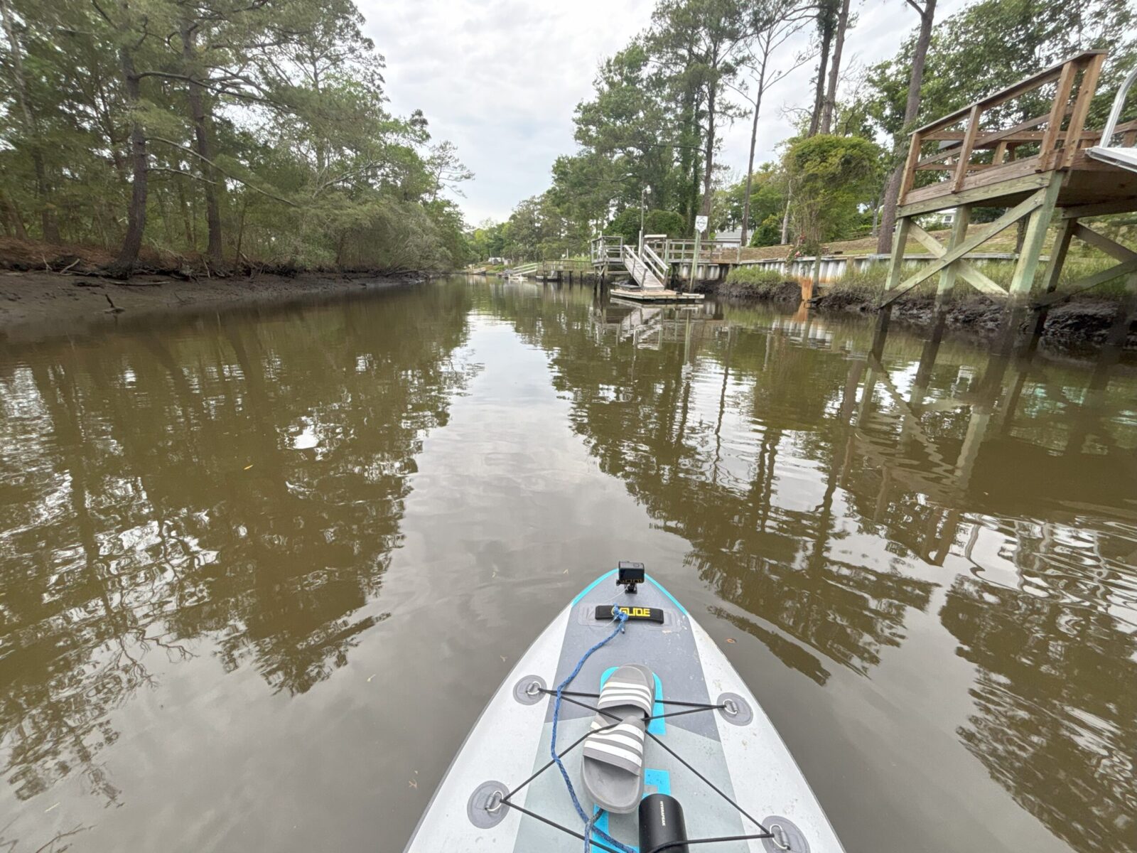 person's POV being on a paddleboard on the oak island canal with brown water, trees and docks nearby.