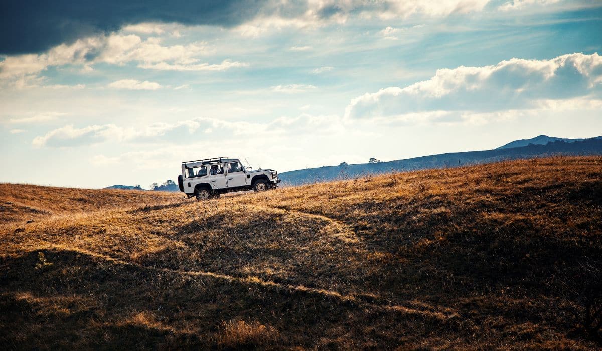 A car driving up a small hill with no road. The sky behind the vehicle is bright and partially cloudy.