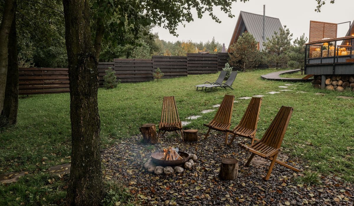 A backyard that has a fire pit with multiple wooden chairs surrounding it. There are also two reclining chairs further back.