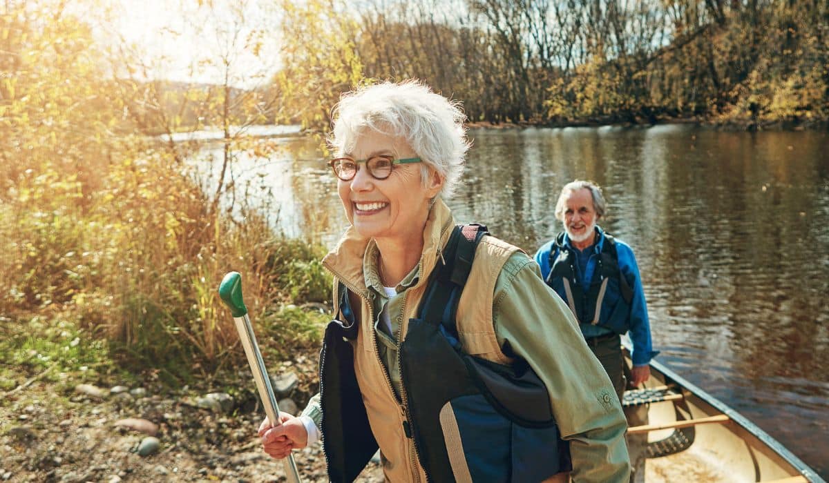 An older woman wearing a life vest smiles while holding a paddle by a lake, with a man and a canoe behind her.