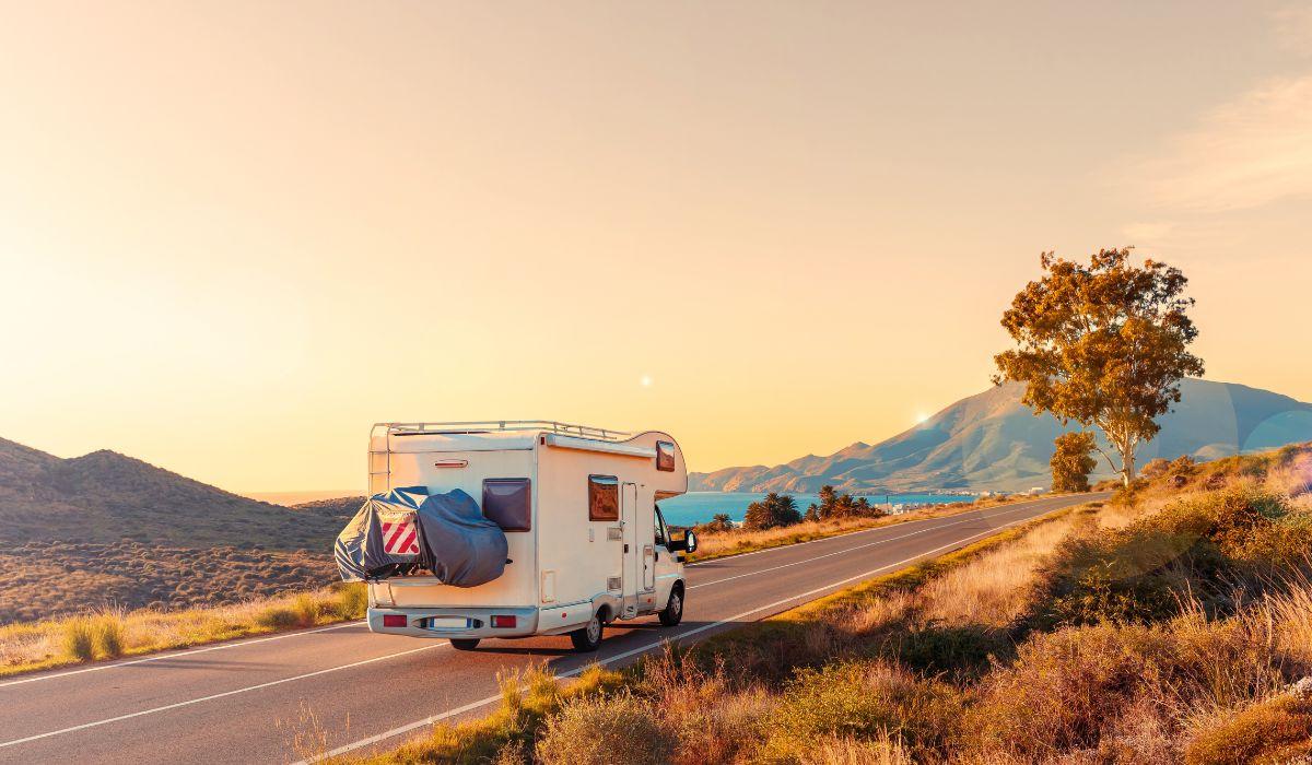 A rear view of a white recreational vehicle camper driving on an empty two-lane road toward a mountain and lake.