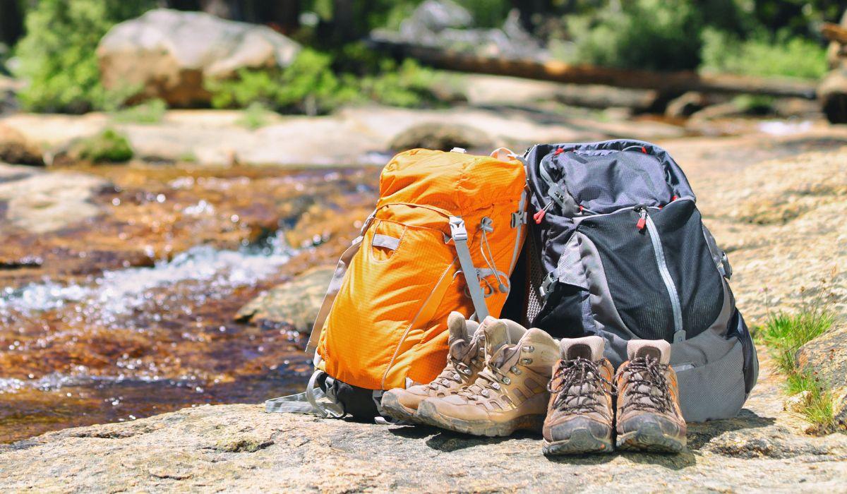 Two hiking backpacks are propped up against each other next to a stream. In the background is a forested area.