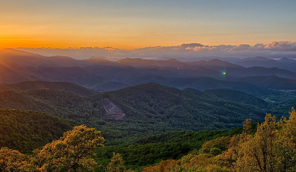 The sun rises over layered Blue Ridge mountains as green forested valleys stretch beneath golden clouds.