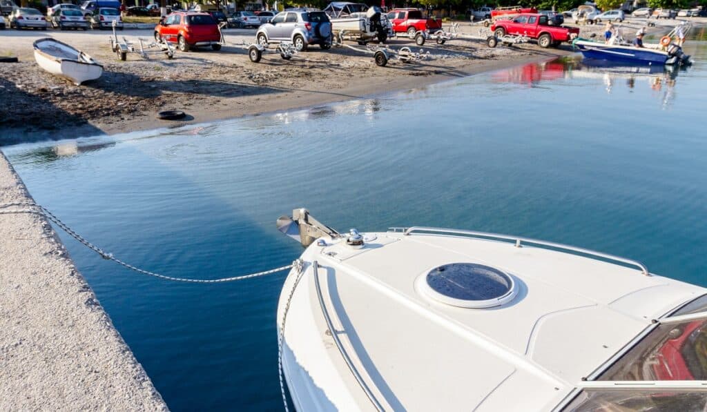 A white boat idles beside a busy launch ramp as trucks and trailers line a muddy lakeside parking area.