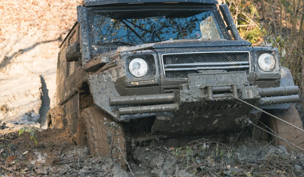 A winch cable pulls a mud-covered SUV through a deep rut on a wooded off-road trail while mud coats the bumper and tires.